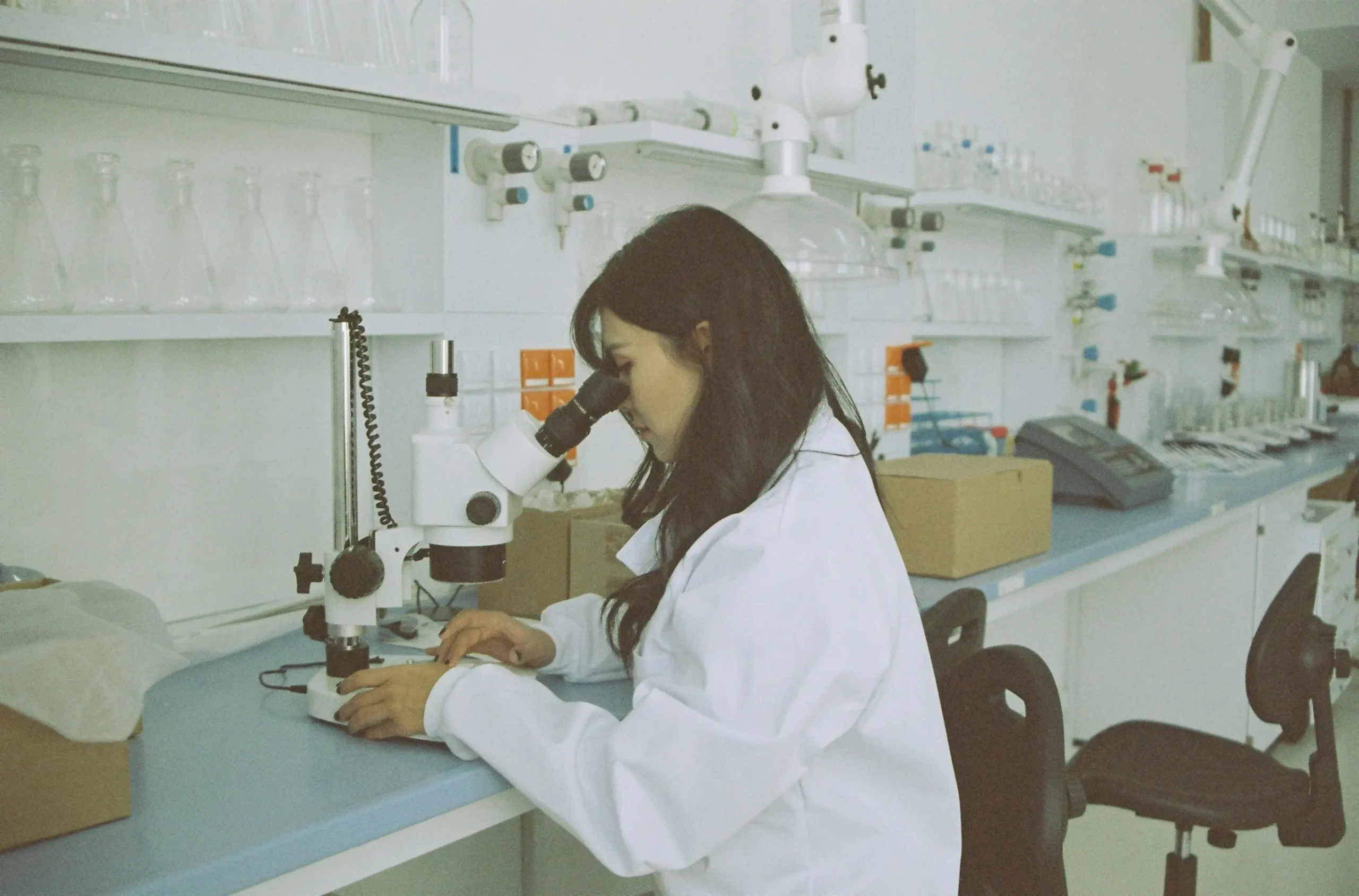 Chemik w fartuchu laboratoryjnym analizujący próbkę pod mikroskopem w laboratorium wyposażonym w szkło laboratoryjne
